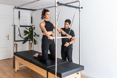 Side view of concentrated female athlete doing exercises on pilates reformer while listening to advice of personal instructor in gym