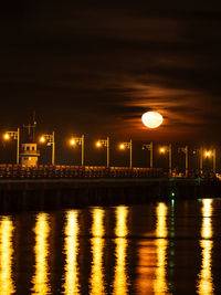 Illuminated street lights by river against sky at night