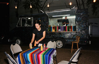 Ethnic female worker putting bottles with sauces on covered table near food truck with male at counter at night