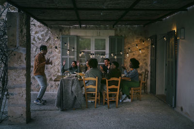 Male and female friends sitting at dining table cheering for man dancing in patio during dinner party