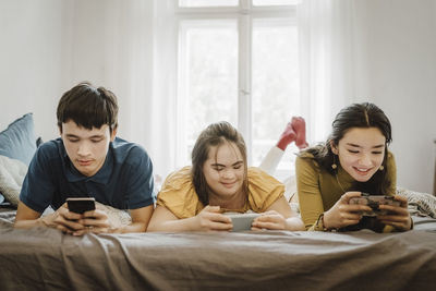 Smiling siblings using smart phones while lying down on bed with down syndrome sister at home