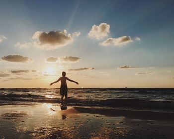 Rear view of shirtless boy with arms outstretched at beach against sky during sunset