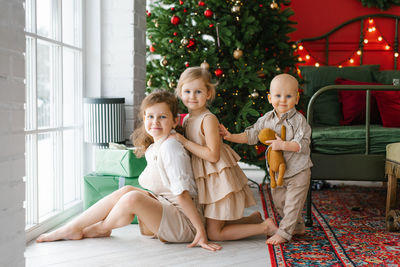 Three children a boy and girls are sitting at the window against the background of a christmas tree 