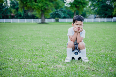 Full length portrait of cute sad boy sitting on soccer ball in park