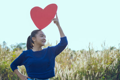 Woman holding heart shape on field against sky