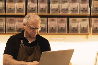 Concentrated mature male barista with gray hair in apron and eyeglasses using laptop while standing at counter in modern coffee shop