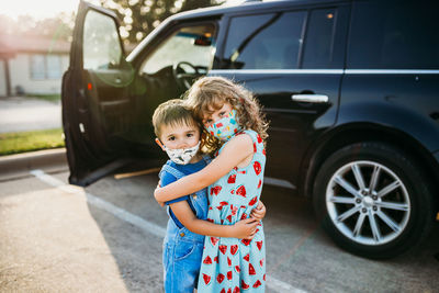 Two young kids standing in parking lot hugging wearing masks