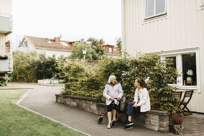 Senior woman and female healthcare worker spending leisure time while sitting on retaining wall against plant