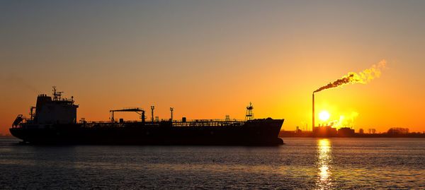 Silhouette factory by sea against orange sky