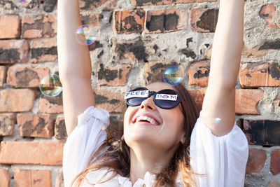 Cheerful woman wearing sunglasses with freedom text while playing with bubbles against brick wall