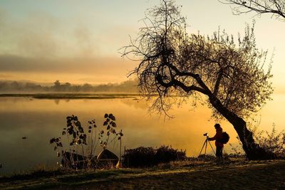 Silhouette man photographing by lake against sky during sunset