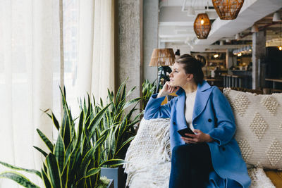 Young woman holding smart phone while looking through window at cafe