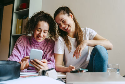 Smiling young women sharing smart phone while sitting with books at home