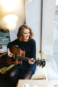 High angle view of smiling young woman playing guitar by window at home