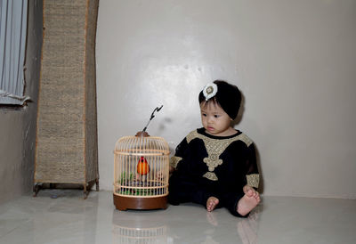 Cute girl looking at bird in cage while sitting on floor at home