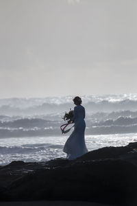 Bride with bouquet standing on shore against sky