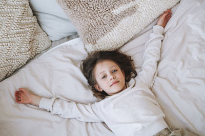 Cute funny little girl with long hair in grey home dress having fun lies on bed at the home