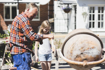 Side view of grandfather using cement mixer with granddaughter in yard