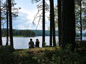 People sitting by tree trunk by lake against sky