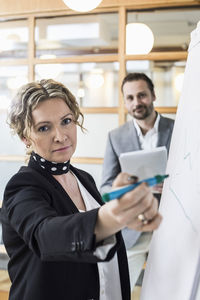 Portrait of mature businesswoman writing on flip chart with male colleague in background at office