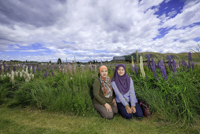 Portrait of smiling mother with daughter kneeling on grassy field against cloudy sky