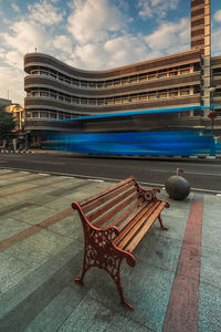 Empty bench by street against buildings in city