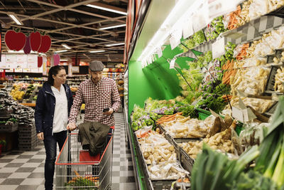 Couple choosing vegetables while using phone in supermarket