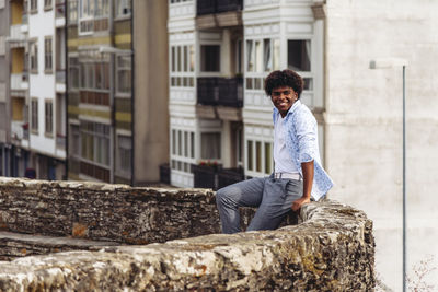 Stylish black male with afro hairstyle sitting on stone on street and smiling while looking away