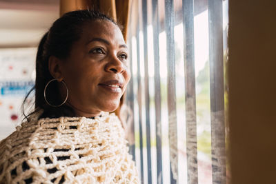 Smiling adult ethnic female standing near window while looking away thoughtfully in light room