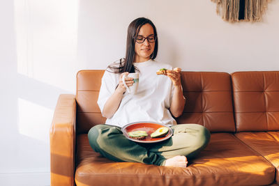 Young modern woman sitting on the sofa drinks coffee and eats a healthy avocado sandwich.