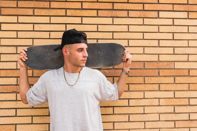 Young man with skateboard standing against brick wall