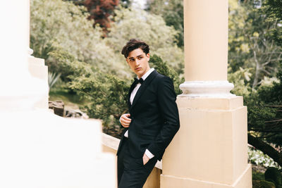 A beautiful young man, the groom in an elegant wedding suit, stands posing in the city's old park