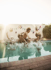 Carefree friends jumping into swimming pool at party against sky during sunset