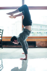 Side view of man practicing handstand on floor against window