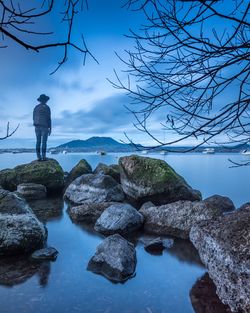 Rear view of woman standing on rock by lake against sky at dusk