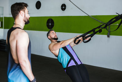 Side view of young muscular man in sportswear training with elastic resistance bands during intense workout with friend in modern gym