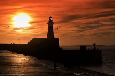 Silhouette lighthouse by sea against orange sky