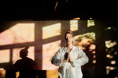 Young mindful male with hands clasped and closed eyes meditating during yoga practice against wall with shades in soft sunlight