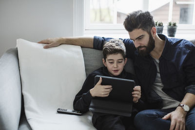 Father looking at boy using digital tablet while sitting on sofa in living room