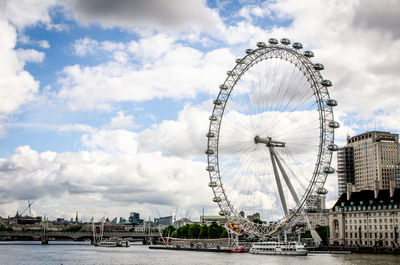 Ferris wheel by river in city against cloudy sky