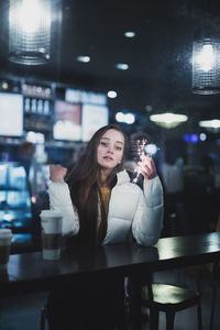 Portrait of young woman holding illuminated string light while sitting at table