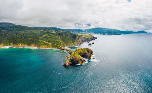 Drone view of paving stone way leading along stone bridge and ridge of rocky hill to lonely house on island gaztelugatxe surrounded by tranquil sea water under cloudy sky in basque country