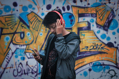 Man listening music while standing against graffiti wall