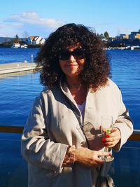 Portrait of woman with curly hair wearing sunglasses against sea in city