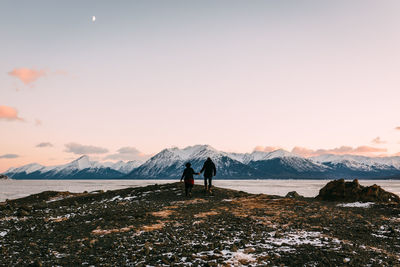 Rear view of people holding hands while walking by lake against mountains and sky during sunset