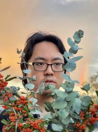 Young asian man with orange rowan berries and eucalyptus leaves against rainbow in sunset sky