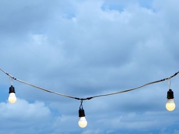 Low angle view of light bulbs hanging against cloudy sky