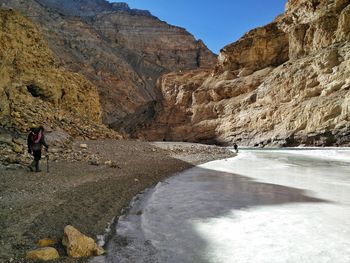 Rear view of man walking along side frozen river against rocky mountains