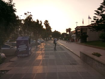 People on footpath by street against sky at sunset