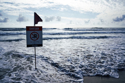 Danger sign board at sea shore against sky